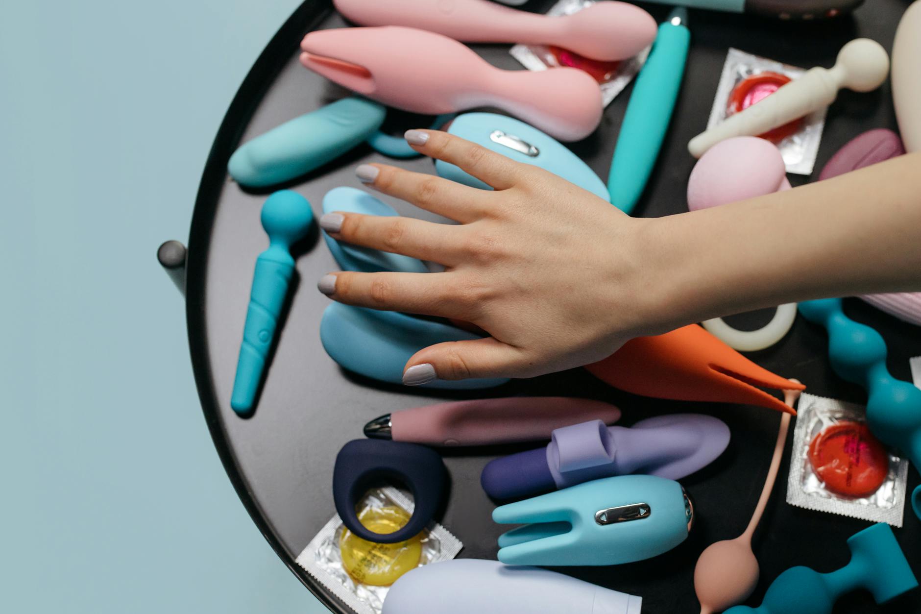 A hand reaching over a variety of colorful clitoral vibrators arranged on a table.