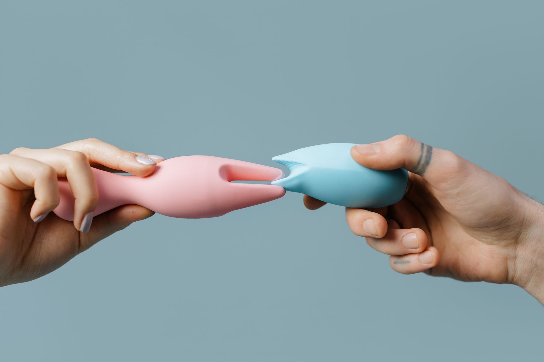 Two hands holding pink and blue silicone vibrators against a pastel background, representing partnership and shared pleasure.